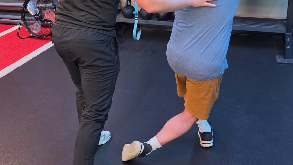 A Physio+ doctor of physical therapy guiding a patient through a curtsy lunge on suspension straps to load the glutes during back pain rehab