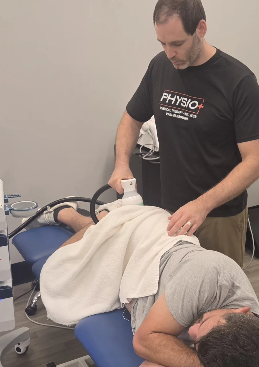 Shockwave being applied to a patient's upper back at Physio+, with the console alongside the treatment table