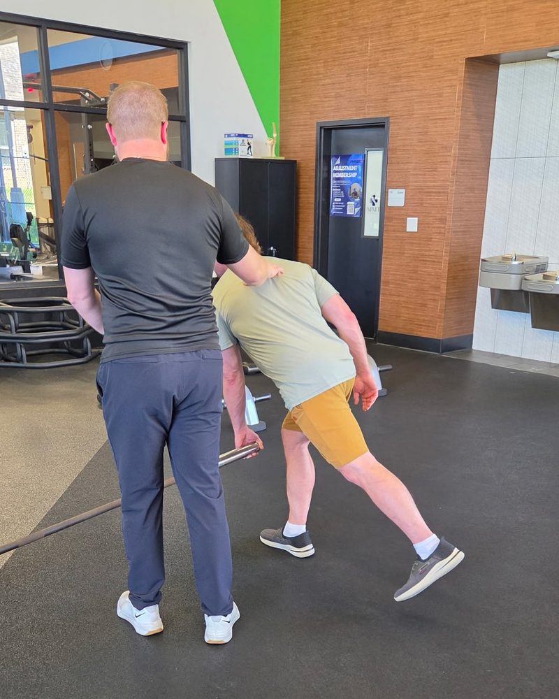 A Physio+ clinician cueing a patient through a single leg Romanian deadlift on a landmine bar